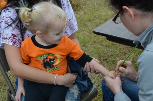A child in an orange shirt sits on a lap while getting a green design painted on their arm.