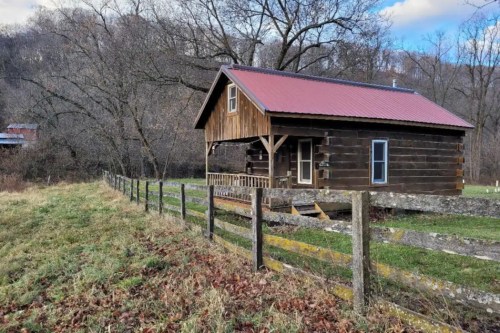 A rustic wooden cabin with a red roof, surrounded by a grassy area and trees, set against a cloudy sky.