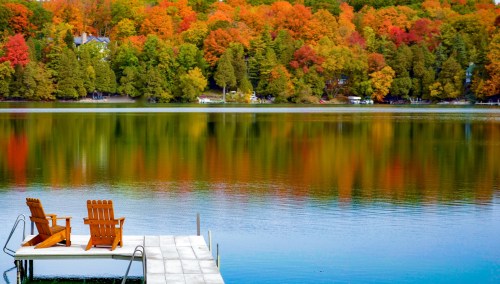 A serene lake view with colorful autumn trees and two wooden chairs on a dock.