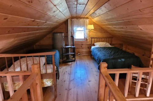 Cozy attic bedroom with wooden walls, two beds, a small table, and a window letting in natural light.