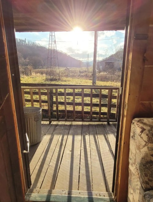 View from a cabin doorway, showcasing sunlight streaming in over a wooden porch and a scenic landscape beyond.