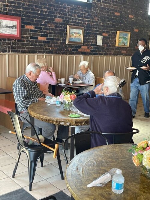 A group of elderly people dining at a cozy restaurant with brick walls and floral centerpieces on the tables.