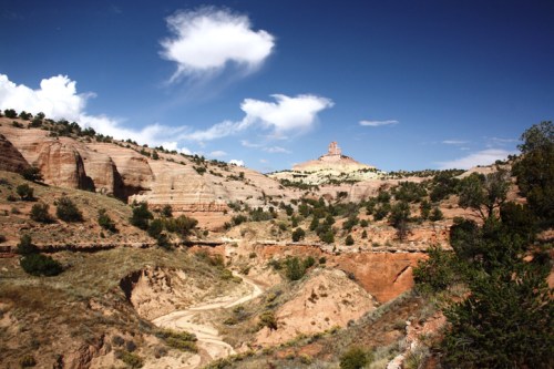 A scenic landscape featuring rocky hills, a winding dirt path, and a prominent rock formation under a blue sky with clouds.