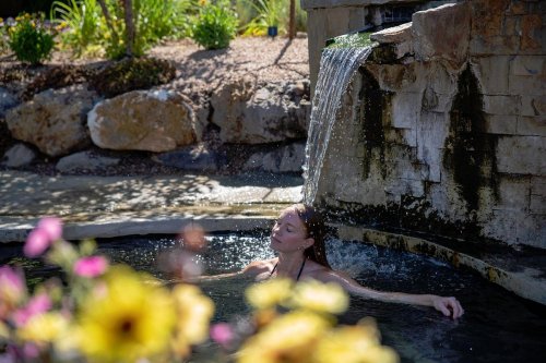 A woman relaxes in a stone pool under a waterfall, surrounded by colorful flowers and greenery.