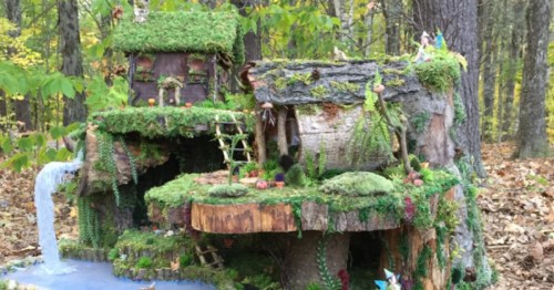 A whimsical fairy house made of wood and moss, featuring a waterfall and surrounded by trees in a forest setting.