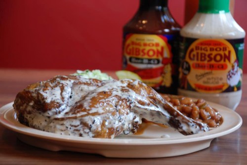A plate of grilled chicken drizzled with white sauce, accompanied by baked beans and barbecue sauces in the background.