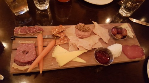 A wooden platter with assorted meats, cheeses, crackers, carrots, and spreads, elegantly arranged for sharing.