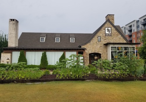 A stone building with a sloped roof, surrounded by greenery and sunflowers, set against a cloudy sky.