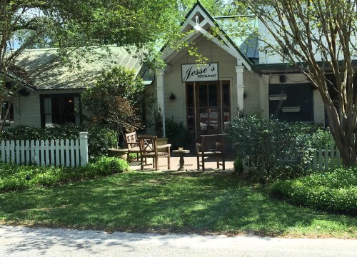 A charming restaurant with a sign reading "Jessie's," featuring outdoor seating and a white picket fence.