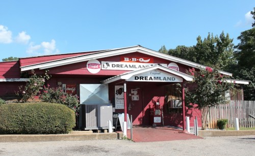 A red building with a sign reading "BBQ Dreamland," featuring outdoor seating and a clear blue sky.