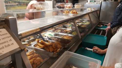 A cafeteria-style food counter with various dishes and customers selecting their meals.