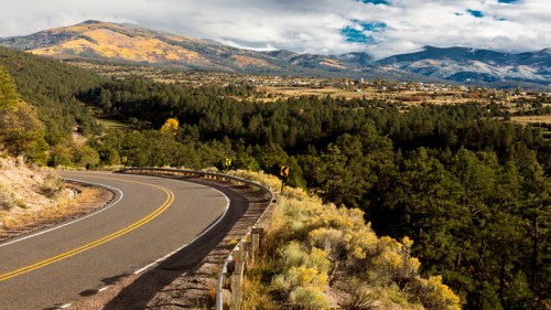 A winding road curves through a mountainous landscape with trees and distant hills under a cloudy sky.