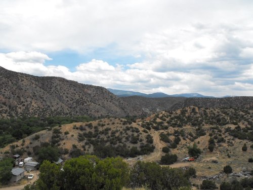 A scenic view of rolling hills and mountains under a cloudy sky, with a small settlement visible in the foreground.