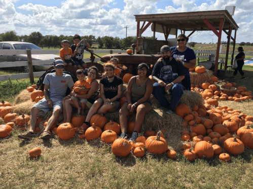 A group of people sitting on hay bales surrounded by pumpkins on a sunny day at a pumpkin patch.