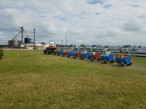 A tractor pulls a colorful train of small carts with children seated, set against a cloudy sky and parked cars.
