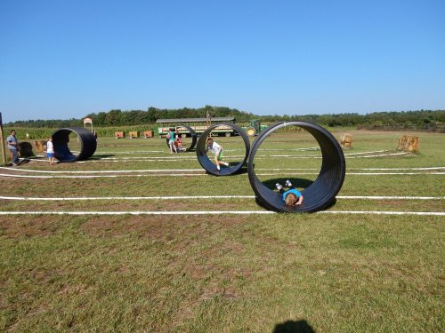Children play in large black tubes on a grassy field, with hay bales and a clear blue sky in the background.