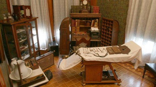 A vintage medical room with an examination table, surgical tools, cabinets, and historical medical equipment.