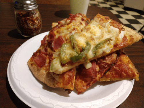 A plate stacked with slices of pizza topped with cheese, pepperoni, and green peppers, alongside a jar of red pepper flakes.
