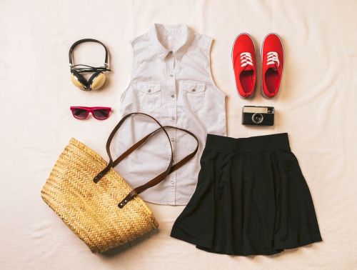 Flat lay of a casual outfit: a sleeveless shirt, black skirt, red sneakers, sunglasses, headphones, camera, and a straw bag.