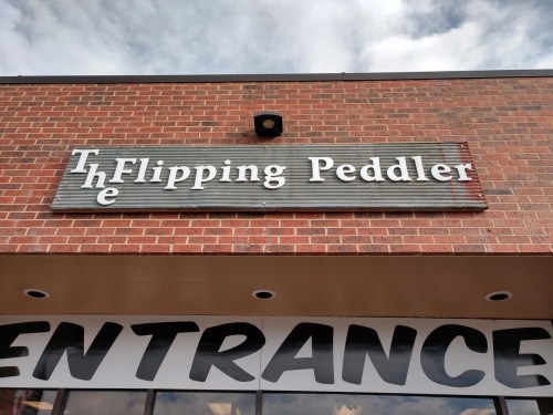 Sign for "The Flipping Peddler" above an entrance, with a brick wall and cloudy sky in the background.