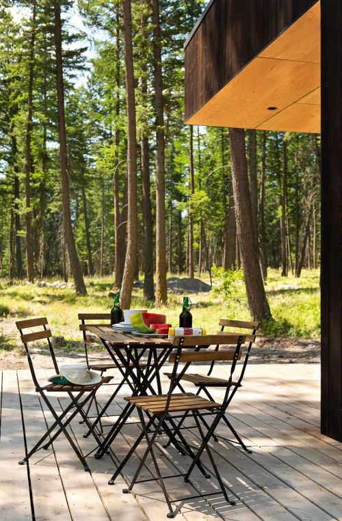 A wooden deck with a table and chairs surrounded by tall trees, featuring drinks and colorful plates.