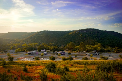 A scenic view of a campground with RVs, surrounded by rolling hills and colorful vegetation under a sunset sky.