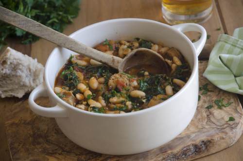 A white pot filled with hearty soup, beans, and greens, with a wooden spoon resting inside, on a wooden surface.