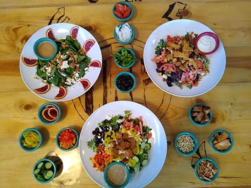 Three colorful salads in large bowls, surrounded by small bowls of toppings and dressings on a wooden table.
