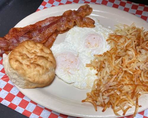 A breakfast plate featuring two fried eggs, crispy bacon, hash browns, and a biscuit on a checkered background.