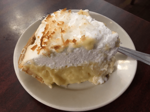 A slice of coconut cream pie with toasted coconut on top, served on a white plate with a fork.