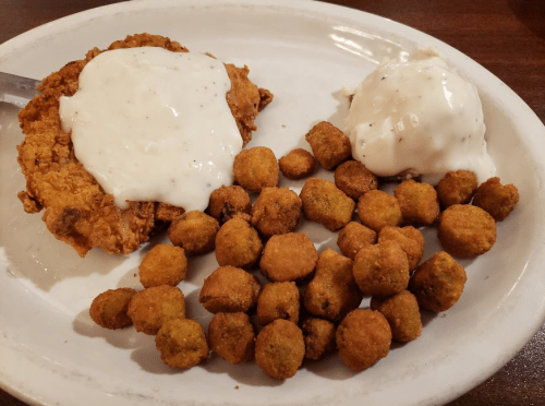 A plate with fried chicken topped with white gravy, served alongside fried okra and a scoop of mashed potatoes.