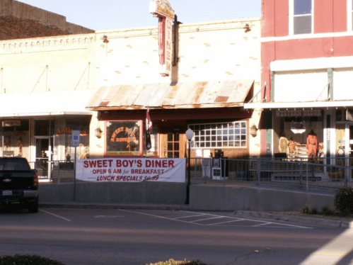 A diner with a sign reading "Sweet Boy's Diner" and hours for breakfast and lunch specials, set in a small town.