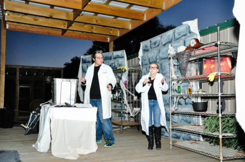 Two people in lab coats perform a science experiment on a stage decorated with spooky props at dusk.