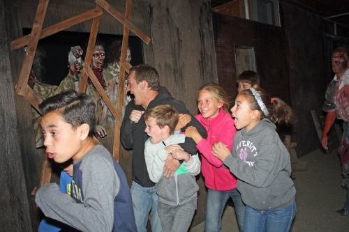A group of children and an adult run in fear past a spooky scene, with zombies visible in the background.