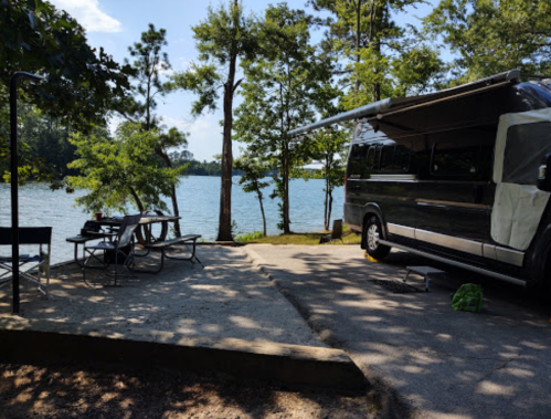 A scenic campsite by a lake featuring a parked RV, picnic table, and trees under a clear blue sky.