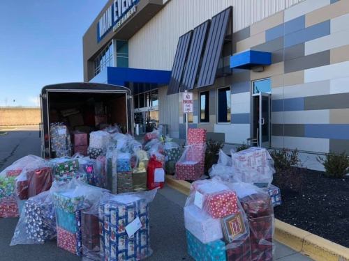 A trailer filled with colorful wrapped gifts outside a building, with many more gifts piled on the ground nearby.