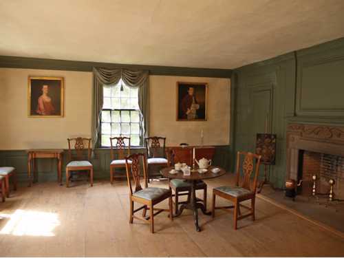 A historic room with green walls, a table set for tea, chairs, and two portraits on the walls.