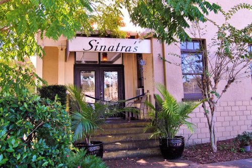 Entrance of Sinatra's restaurant, featuring a sign, steps, and potted plants surrounded by greenery.