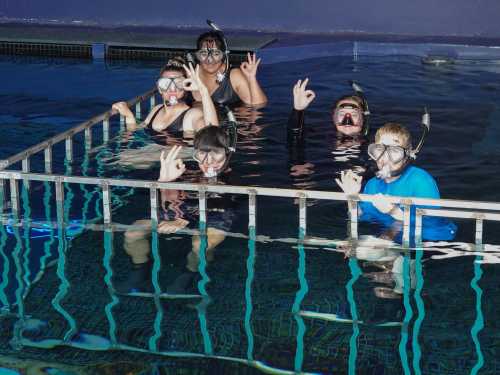 A group of five people in snorkeling gear smiles and poses in a pool, with water reflections visible.