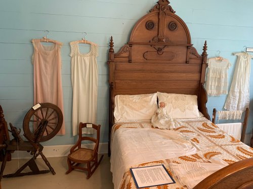 A vintage bedroom with a wooden bed, a doll on the bed, and antique clothing displayed on the wall.