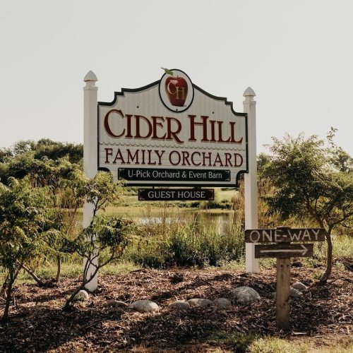 Sign for Cider Hill Family Orchard with U-Pick and Event Barn, surrounded by greenery and a one-way sign nearby.