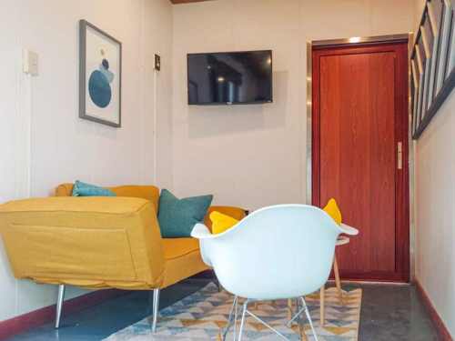 Cozy living space featuring a yellow couch, white chair, TV, and a wooden door. Decorated with colorful cushions and art.