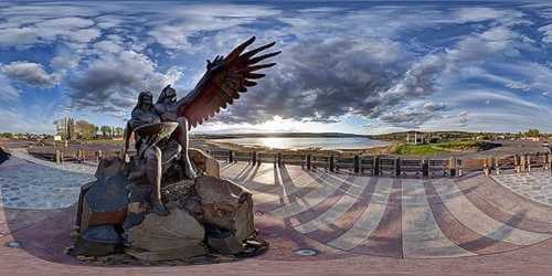 A panoramic view of a bronze statue with wings, set against a scenic lake and cloudy sky.