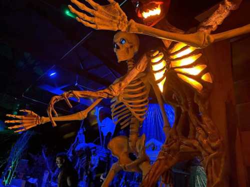 A large skeleton figure with glowing eyes stands in a dark, decorated space, surrounded by eerie Halloween decorations.