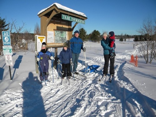 A family stands by a trail information sign in a snowy landscape, ready for cross-country skiing.