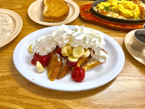 A plate of French toast topped with whipped cream, bananas, and strawberries, with toast and a skillet meal in the background.