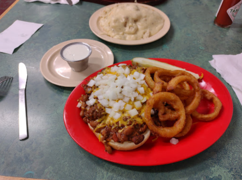 A red plate with a loaded hot dog topped with onions and mustard, served with onion rings and a side of ranch dressing.