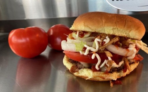 A close-up of a burger with lettuce, tomato, and toppings, alongside two fresh tomatoes on a metal surface.