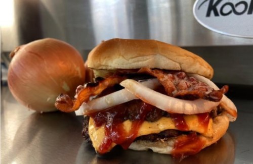 A juicy burger with bacon, cheese, and onions, alongside a whole onion on a stainless steel surface.