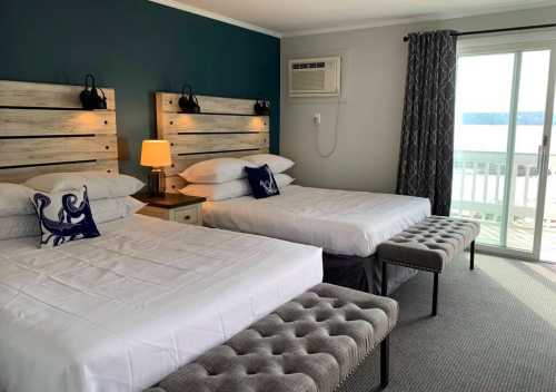 A cozy hotel room with two beds, a bench, and a view of the water through a sliding glass door.